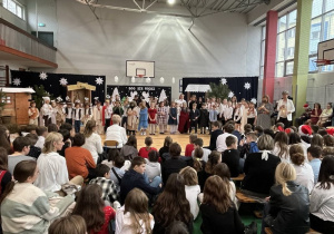 Panie wicedyrektor Małgorzata Lisiak oraz Barbara Duda składają uczniom życzenia stojąc na tle artystów wystawiających szkolne jasełka.