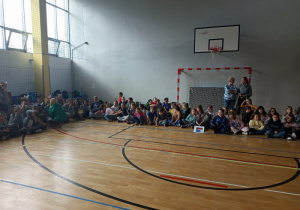Uczniowie siedzą skrzyżnie i obserwują występy poszczególnych klas.