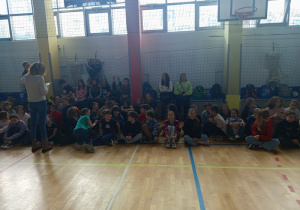 Uczniowie siedzą skrzyżnie i obserwują występy poszczególnych klas.