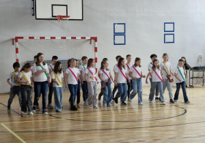 Uczniowie klasy 2c, przepasani kolorowymi szarfami, śpiewają piosenkę „Krajka”