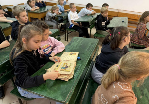 Dzieci siedzą w dawnych ławkach szkolnych i przeglądają stare wydania elementarzy.