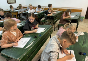 Dzieci siedząc w ławkach piszą gęsim piórem maczanym w kałamarzu.