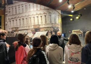 Uczniowie przebywają na parterze Muzeum Powstania, słuchają przewodnika, który opowiada o czasie okupacji i wybuchu Powstania.