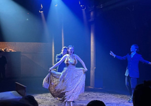Grupa Aktorów występuje na scenie teatralnej wypełnionej ziemią.