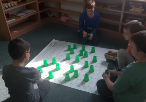 Uczniowie klasy III do kodowania na dywanie wykorzystują plansze i kolorowe, plastikowe kubeczki.
