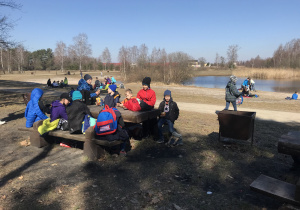 Uczniowie siedzą w lesie przy drewnianych stołach, odpoczywają, jedzą drugie śniadanie.