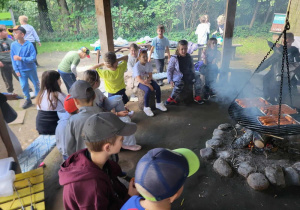 Dzieci siedzą przy ognisku i czekają na piekące się kiełbaski.