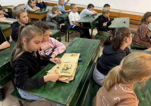 Dzieci siedzą w dawnych ławkach szkolnych i przeglądają stare wydania elementarzy.