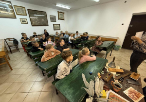 Uczniowie siedząc w ławkach słuchają opowieści o szkole sprzed lat.