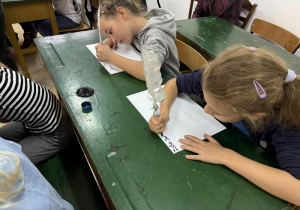 Dzieci siedząc w ławkach piszą gęsim piórem maczanym w kałamarzu.