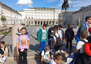 Dzieci podczas spaceru po Warszawie słuchają Pana przewodnika opowiadającego im o Pałacu Prezydenckim.