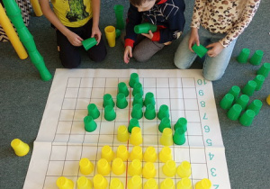 Uczniowie siedzący na dywanie przy macie do kodowania , na której ułożony jest żółty tulipan z kubeczków.