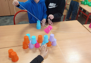 Dzieci za pomocą gumki recepturki ze sznurkami budują wieżę z kubeczków .