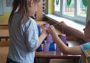 Dzieci za pomocą gumki recepturki ze sznurkami budują wieżę z kubeczków .