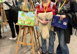 Uczennice z klasy 7b stoją przy swojej pracy konkursowej zaprezentowanej w hali EXPO.