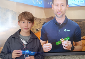 Uczeń z klasy 5d stoi z „kluczem do kosmosu”. W drugim tle widnieje postać Sławosza Uznańskiego - Wiśniewskiego – polskiego inżyniera, astronauty Europejskiej Agencji Kosmicznej.