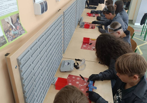Uczestnicy warsztatów wraz z asystentami z koła „Laboratorium Przyszłości” poznają elementy zestawu, w tym płytkę PCB w kształcie satelity oraz komponenty elektroniczne.